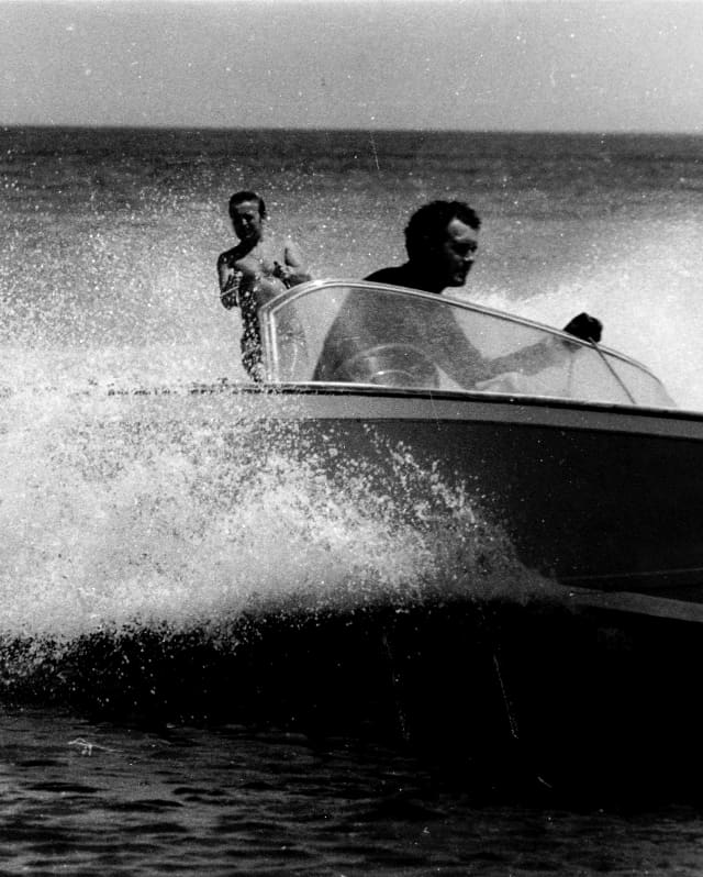 Historic black and white image of a man steering a speed boat, kicking up spray as he tows a water-skier in his wake.