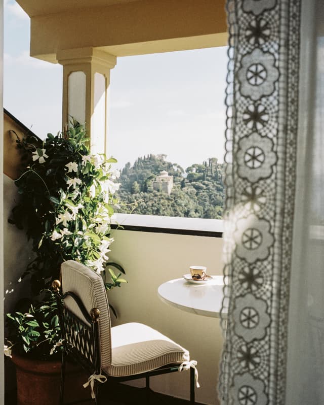 A coffee sits on a table in a warm terrace corner with a chair and blooming orange blossom, seen from behind a lace drape.