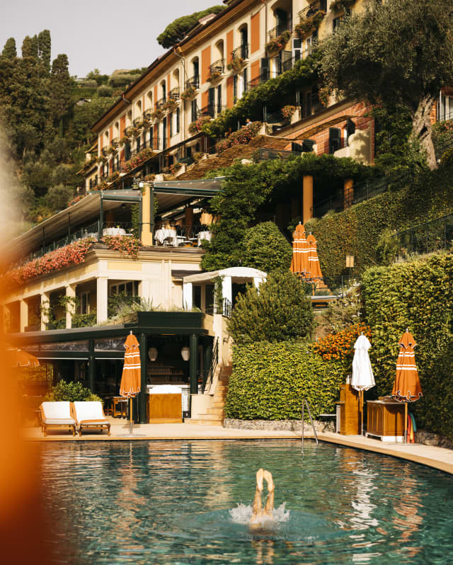 A swimmer performs an underwater handstand in the cobalt pool, with a hotel backdrop of saturated terracotta and cream.