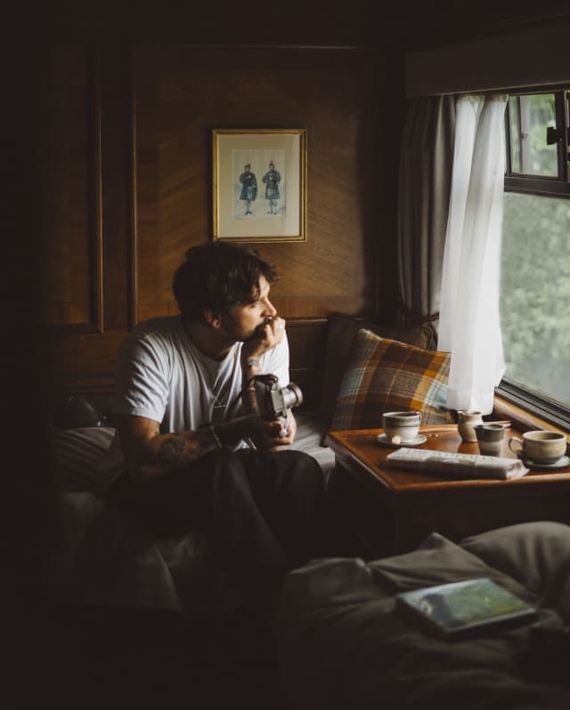 Holding a camera, a man sits with his chin on his hand and his elbow on his knee, looking pensively through his cabin window.