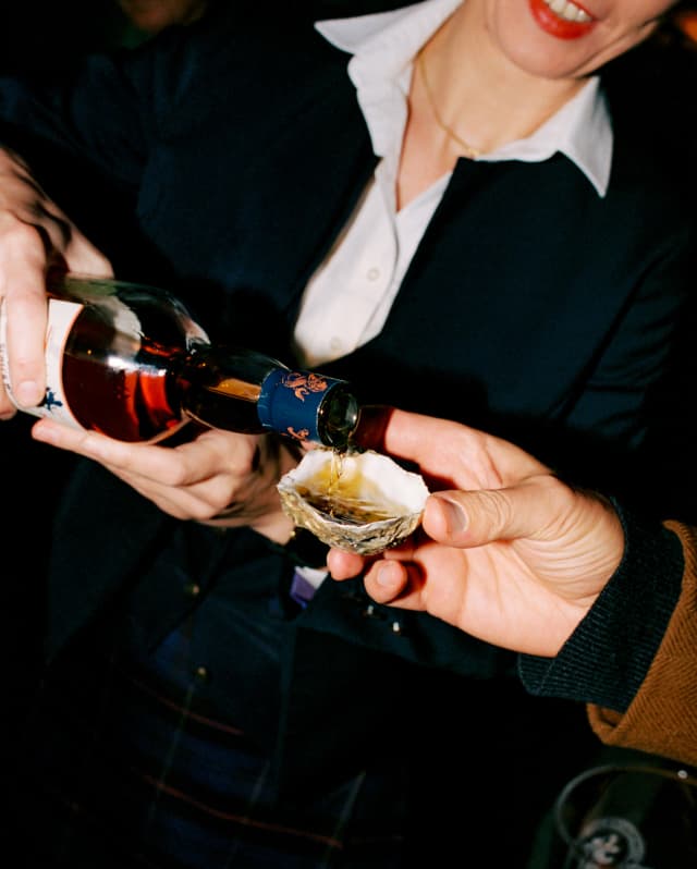 A man, out of shot, holds an oyster shell while a woman in black top over a white shirt pours in whisky, in an angled image.
