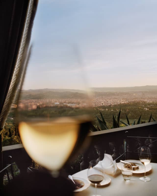 A close up image of a glass of white wine fades into a long view of Florence, its white walls and red roofs surrounded by green