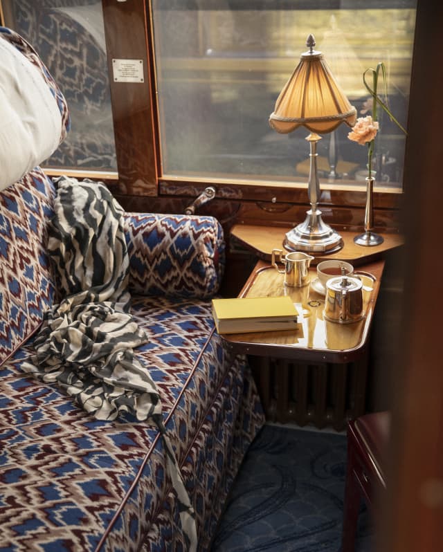 A lamp, a pot of tea and a book rest on a table in a cabin interior, with upholstered banquette in rich blues and maroons.