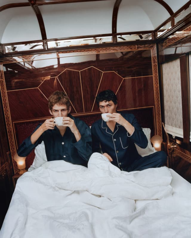 Two men in navy pyjamas sip coffee beneath the white duvet of their bed in the Prague Suite, photographed by Coco Capitán.