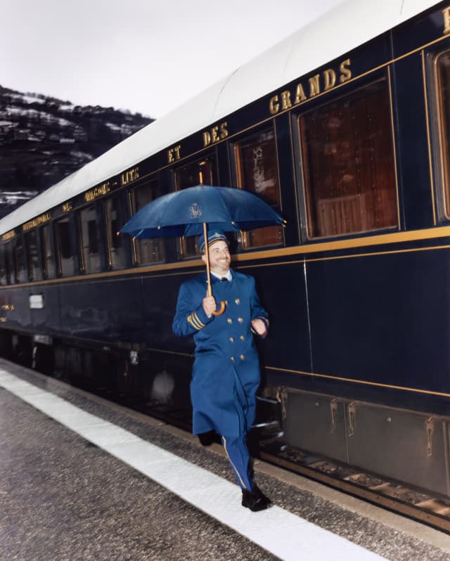 A steward in blue coat with gold buttons and cuff stripes smiles as he dashes through the rain below a matching blue umbrella