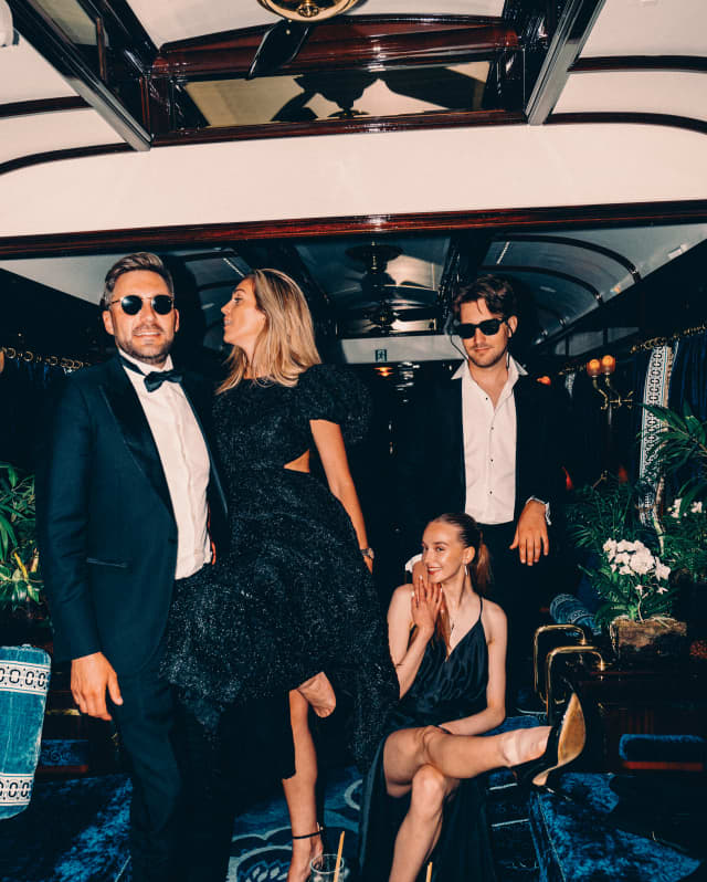 There is a sense of carousing in the air as two men in black tie and sun glasses laugh with two women in black cocktail dresses