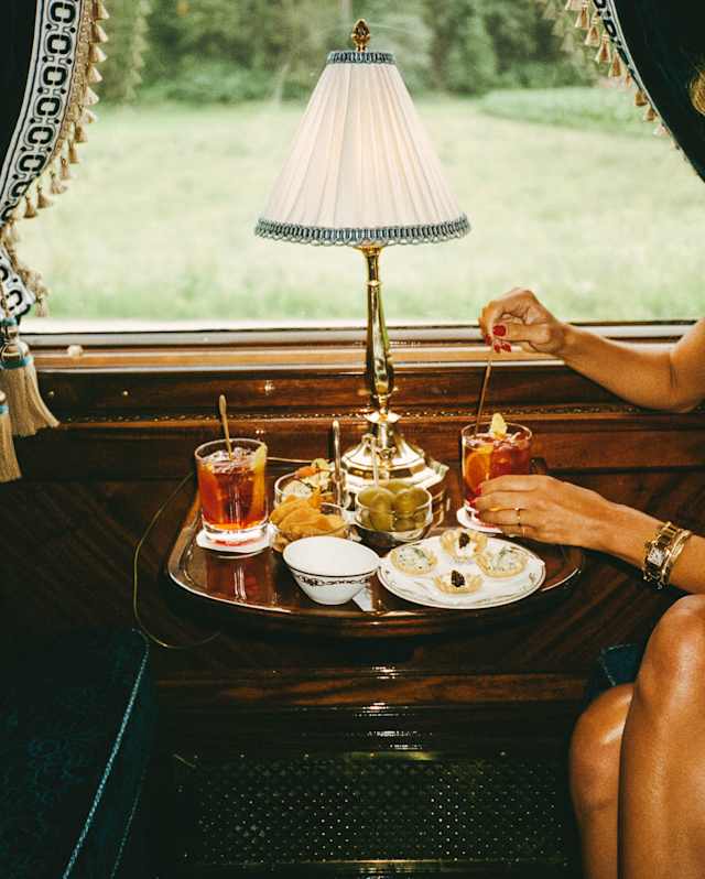 In the Bar Car, a woman with bare legs, mainly off-camera, stirs a Negroni cocktail at a table with aperitifs and nibbles.
