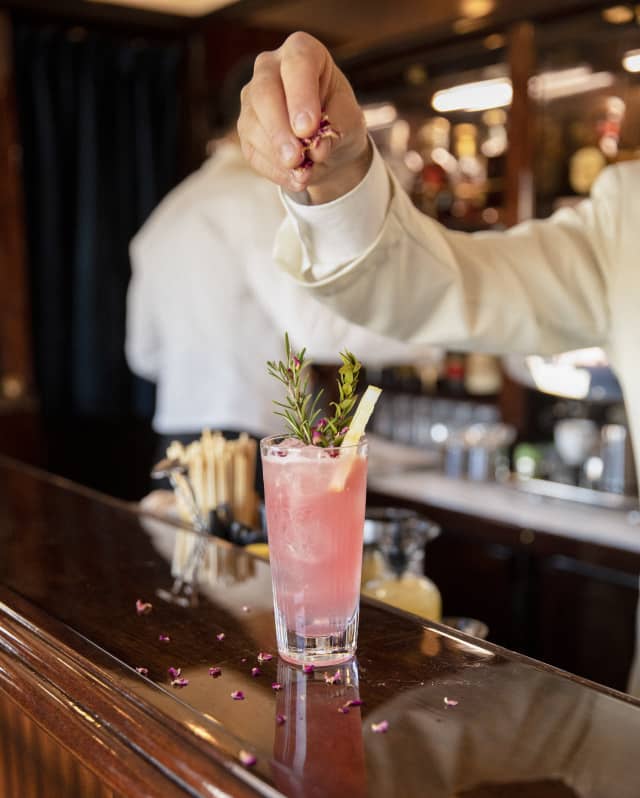 A barman's fingers scatter petal garnish on a grapefruit-pink cocktail with a rosemary sprig, on the gleaming wood bar top.