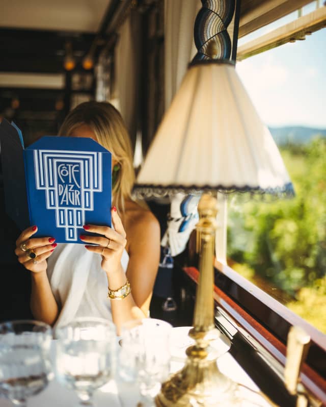 Seen across a table with a lamp and three glasses, a woman holds a blue Cote d'Azur Dining Car menu in front of her face.