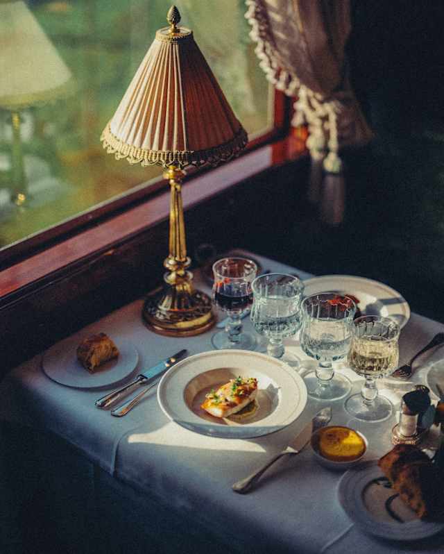 A shaft of sunlight through a window falls onto a gourmet plate of white fish at a dining table with wine and water glasses.