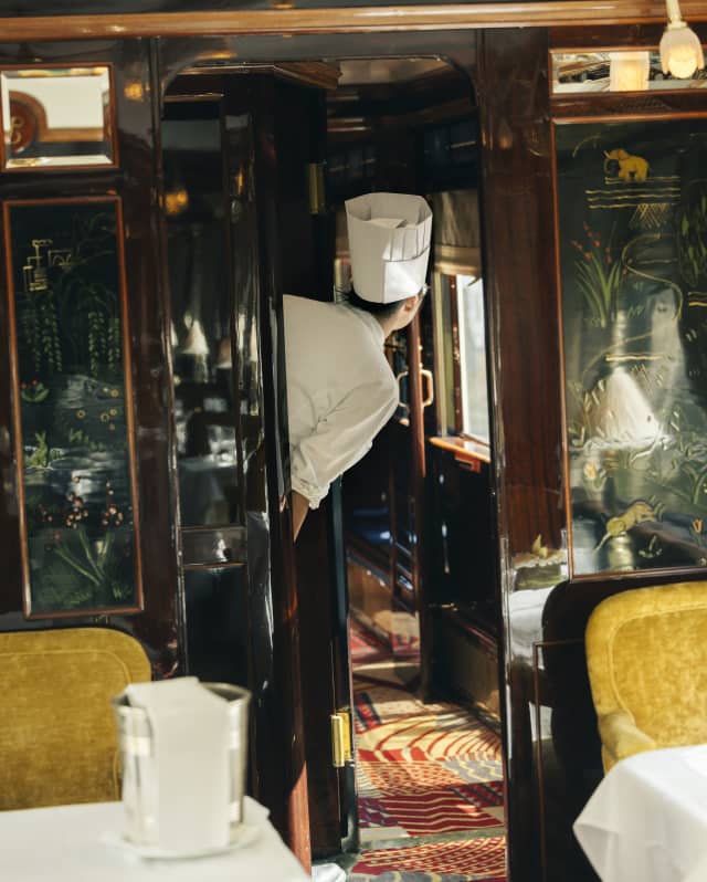 A chef in a toque leans through a doorway in the L'Oriental dining car with yellow chairs and a carpet in sunset shades.