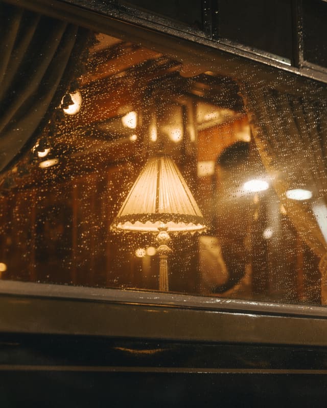 The inviting warmth of a table lamp in the dining car glows through the rain-spattered train window, seen from outside.