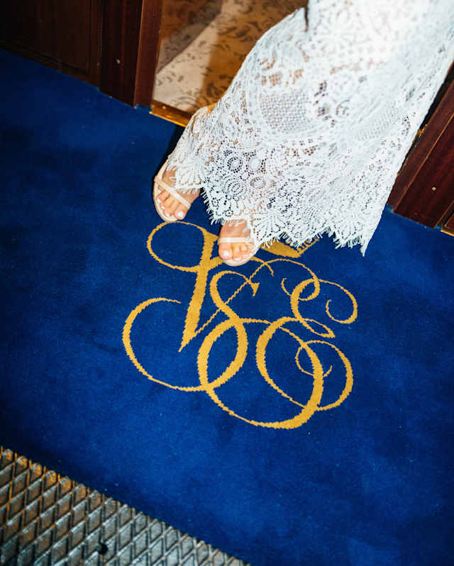Seen from the waist down, a woman in a white lace dress and heels stands on a blue velvet carpet with the gold VSOE monogram.