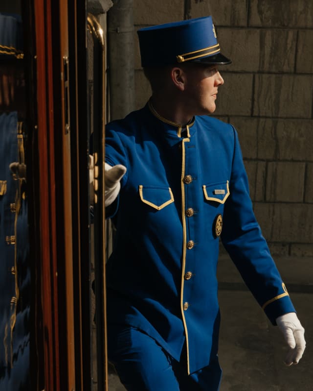 A man in a blue Pullman Porter uniform with gold braiding and a kepi hat stands in the doorway of the stationary train.