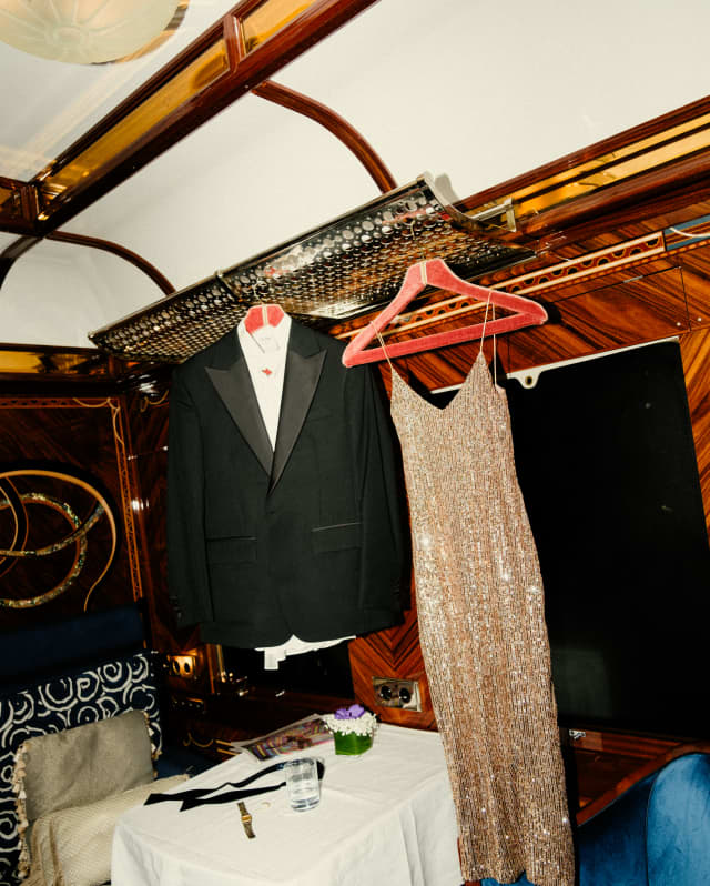 A sparkling gold evening dress and a tuxedo dangle from coat hangers above a table and a blue velvet chair in a cabin.