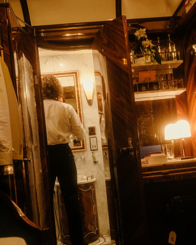 A man in black trousers and white shirt checks his tie in the bathroom mirror, seen from the warm-lit cabin interior behind.