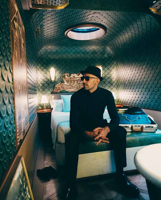 Artist JR, in black clothes, hat and shades sits on the end of a bed in the leather-tiled cabin, gazing at a marquetry panel.