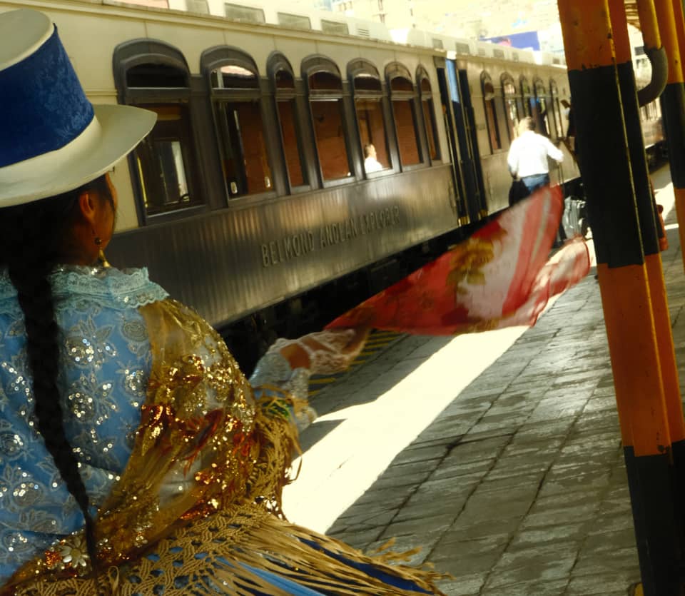 Seen from behind, a local dancer in a glittering gold shawl and white fedora hat welcomes the train at the platform.