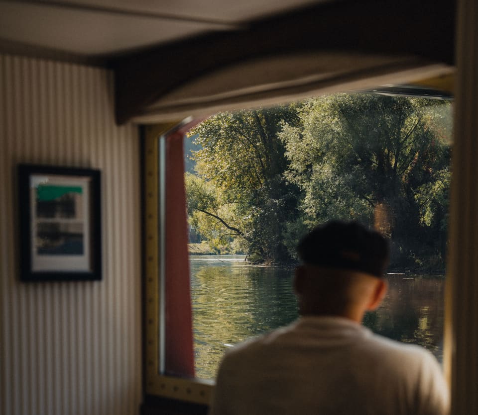 A male guest in a white top and black flat-cap watches the tree-lined waterway from the lounge window, seen from behind.