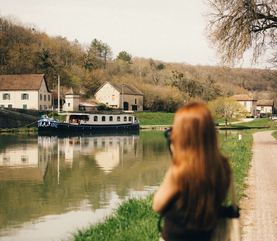 A woman stands on the bank of the canal, taking a photograph of the Fleur de Lys as it cruises by a cluster of white houses.