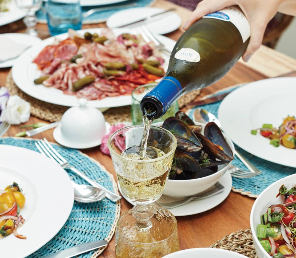 White wine is poured into a crystal glass as guests enjoy a feast, with plates of charcuterie, lentils, salad and shellfish.