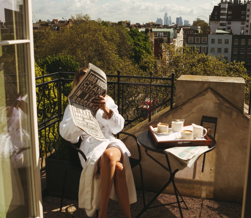 A female guest in a white bath robe reads a newspaper at a table set for morning coffee on a balcony with big views.