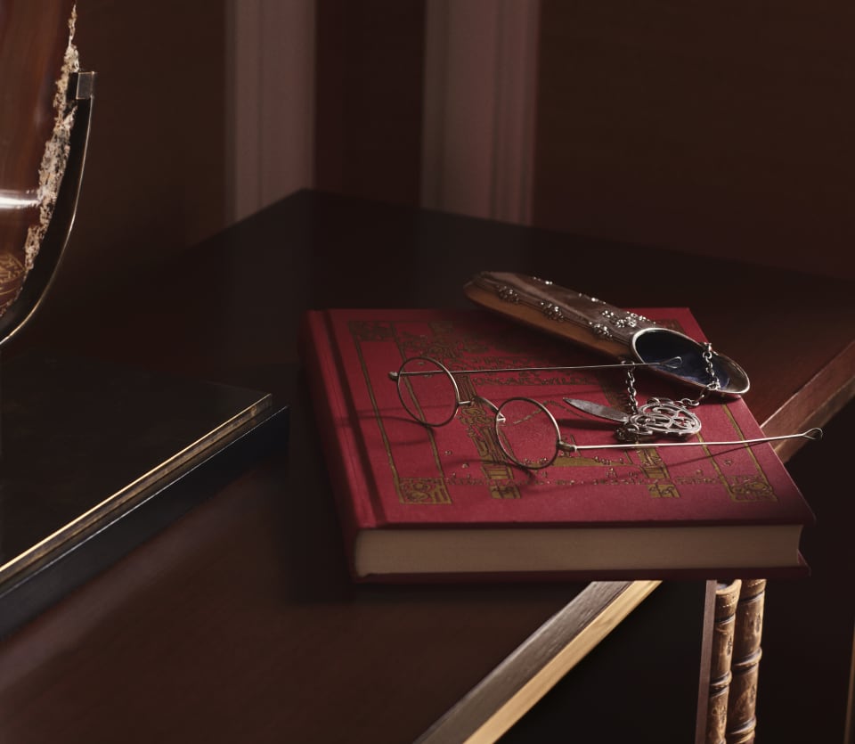 Round, metal-framed spectacles rest on the top of a red, hardback book along with a glasses case in a Penthouse Suite detail.