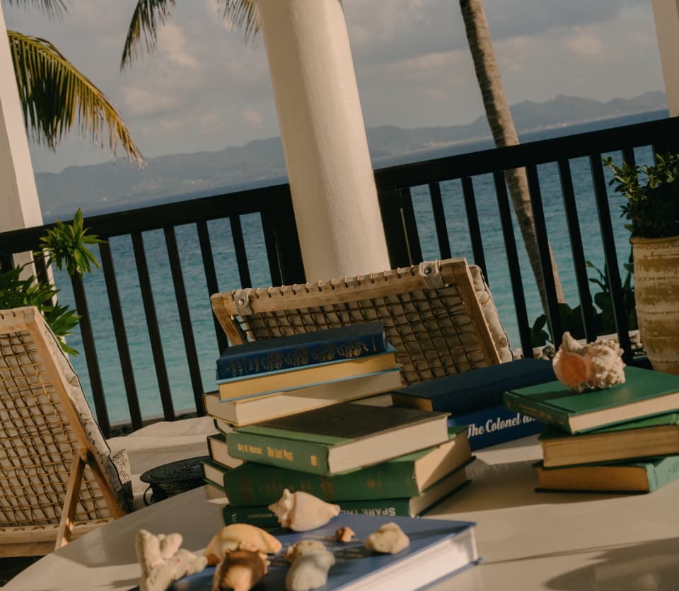 Angled shot of stacked books and seashells on the table of a Pool Villa balcony with sapphire sea views through white arches.