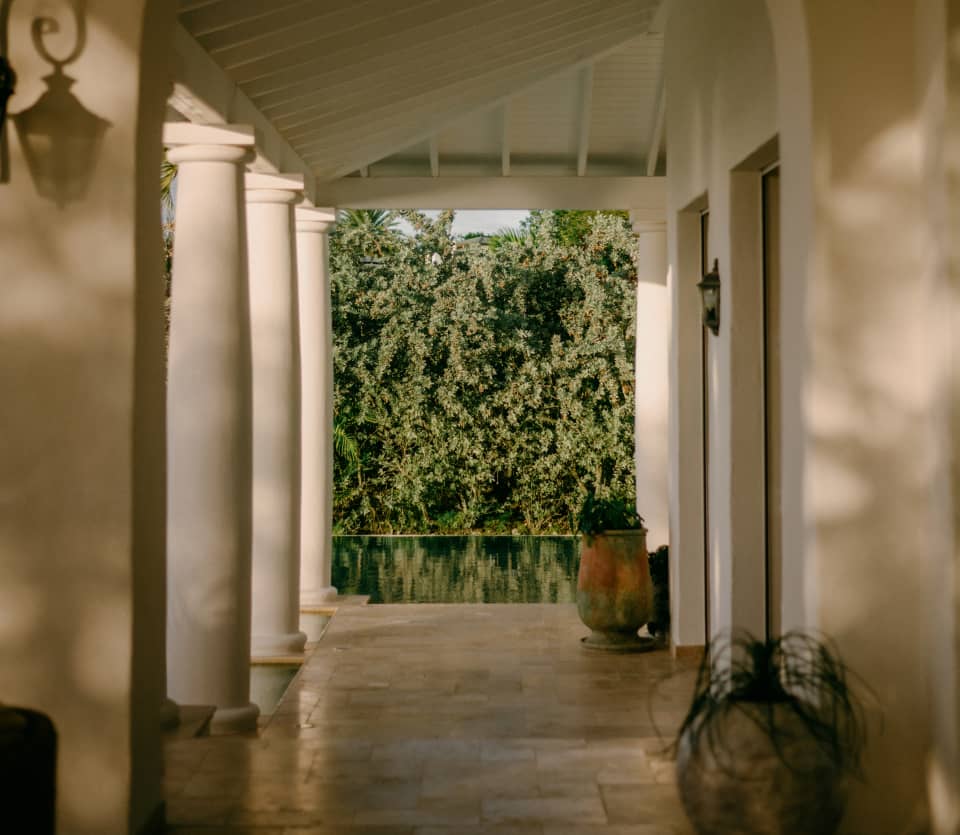 View through the cool, tiled corridor of a veranda, supported by white columns, to the adjacent pool, which reflects foliage.