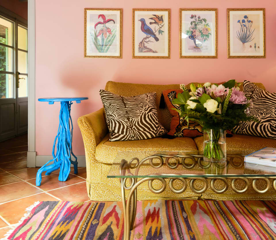 Eight flora and fauna prints adorn the wall above a bronze sofa with print cushions, seen over a glass table on a bright rug.