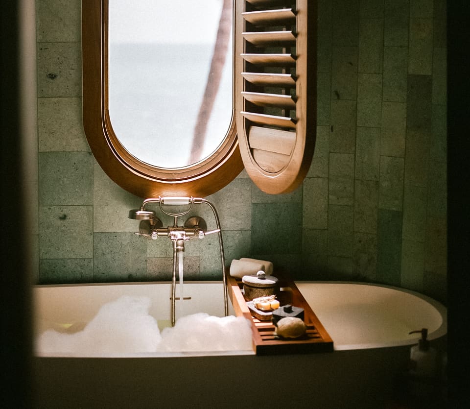 A shutter opens to reveal sea views through an oval window set in the wall above a bubbling bath with a cross-tub soap tray.