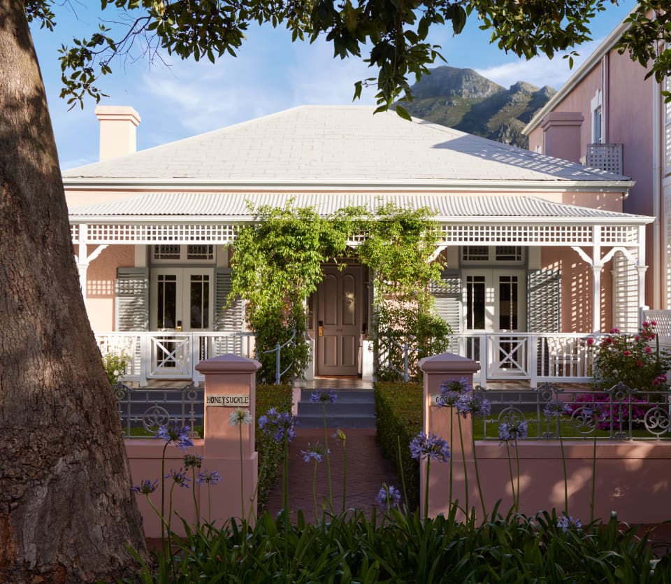 View of the quaint, double-fronted Honeysuckle Cottage with pink garden wall and trellis-like awnings, climbing with vines.