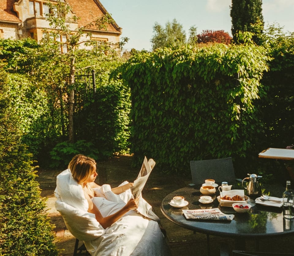 A woman wrapped in a white duvet reads a paper in a secluded garden. Before her the table is laden with breakfast