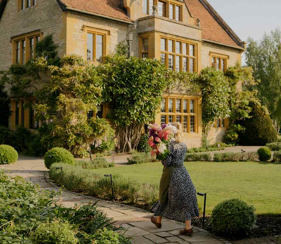 A florist in a patterned dress carries a large bunch of pink garden flowers on her shoulder as she walks towards the house.