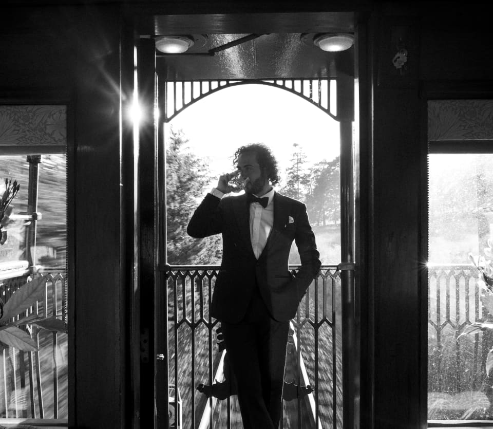 In black and white, a male guest in black-tie sips a drink with one hand in his pocket on the open-air observation deck.